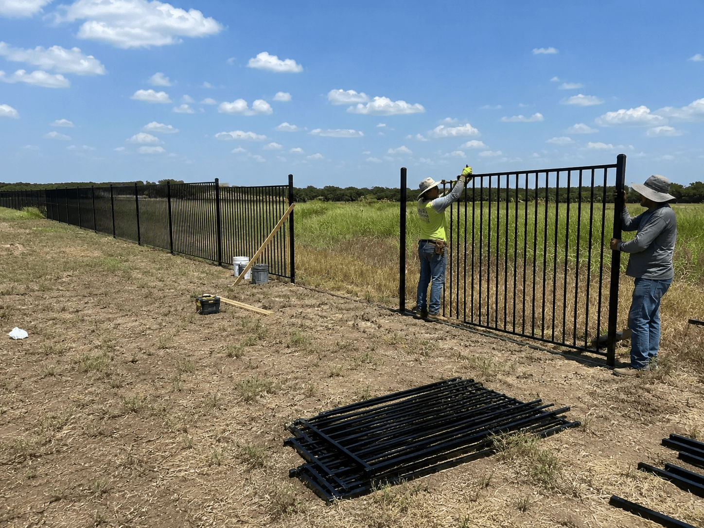 Two person crew setting a black ornamental steel fence panel along a North Texas pasture line, with the completed run stretching back toward the tree line